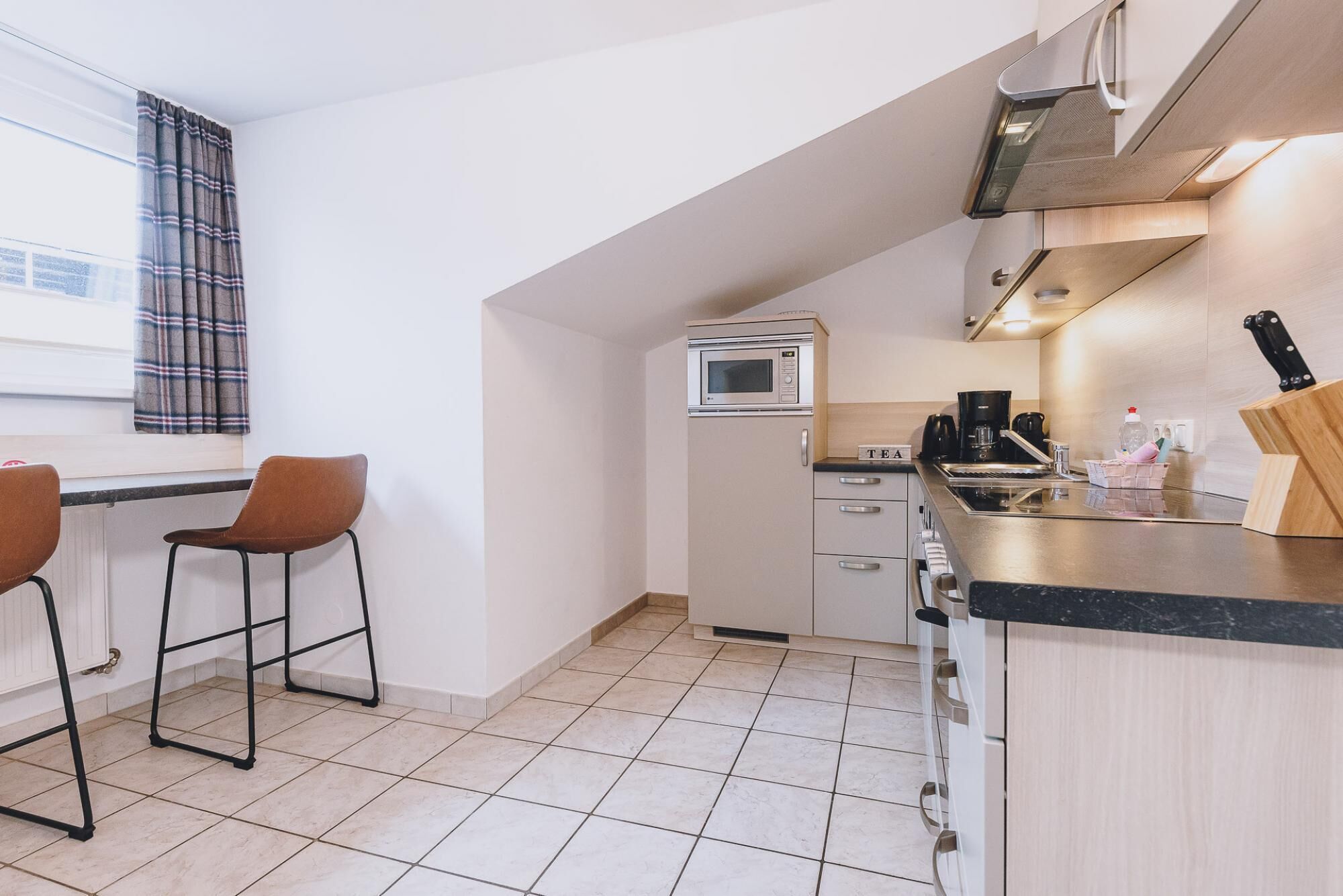 A compact kitchen with a microwave, coffee maker, and knife block. There are two bar stools and a window with curtains.