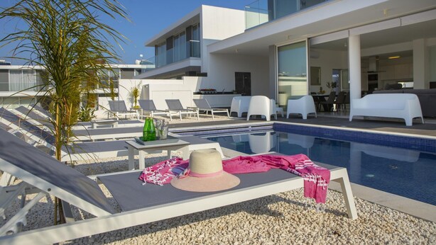 A modern pool area with lounge chairs, a pool, and a building with large windows. - Protaras Olivine Pearl Villa 16 (Paralimni)
