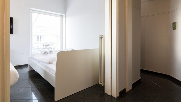 Quarto de hotel moderno com uma janela grande, uma cama, um radiador e uma porta que dá para outro quarto.