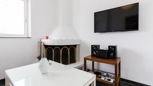 Uma sala de estar moderna com lareira branca, uma TV de tela plana na parede e uma mesinha de madeira com caixas de som.