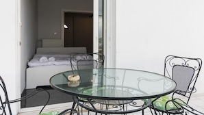 Uma mesa com tampo de vidro e cadeiras de metal, uma cama com roupa de cama branca e um radiador ao fundo.