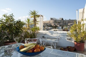 A rooftop terrace with a table holding a bowl of fruit, potted plants, and a view of ancient ruins. - Modern - Vintage Apartment by Acropolis Museum (Athens)