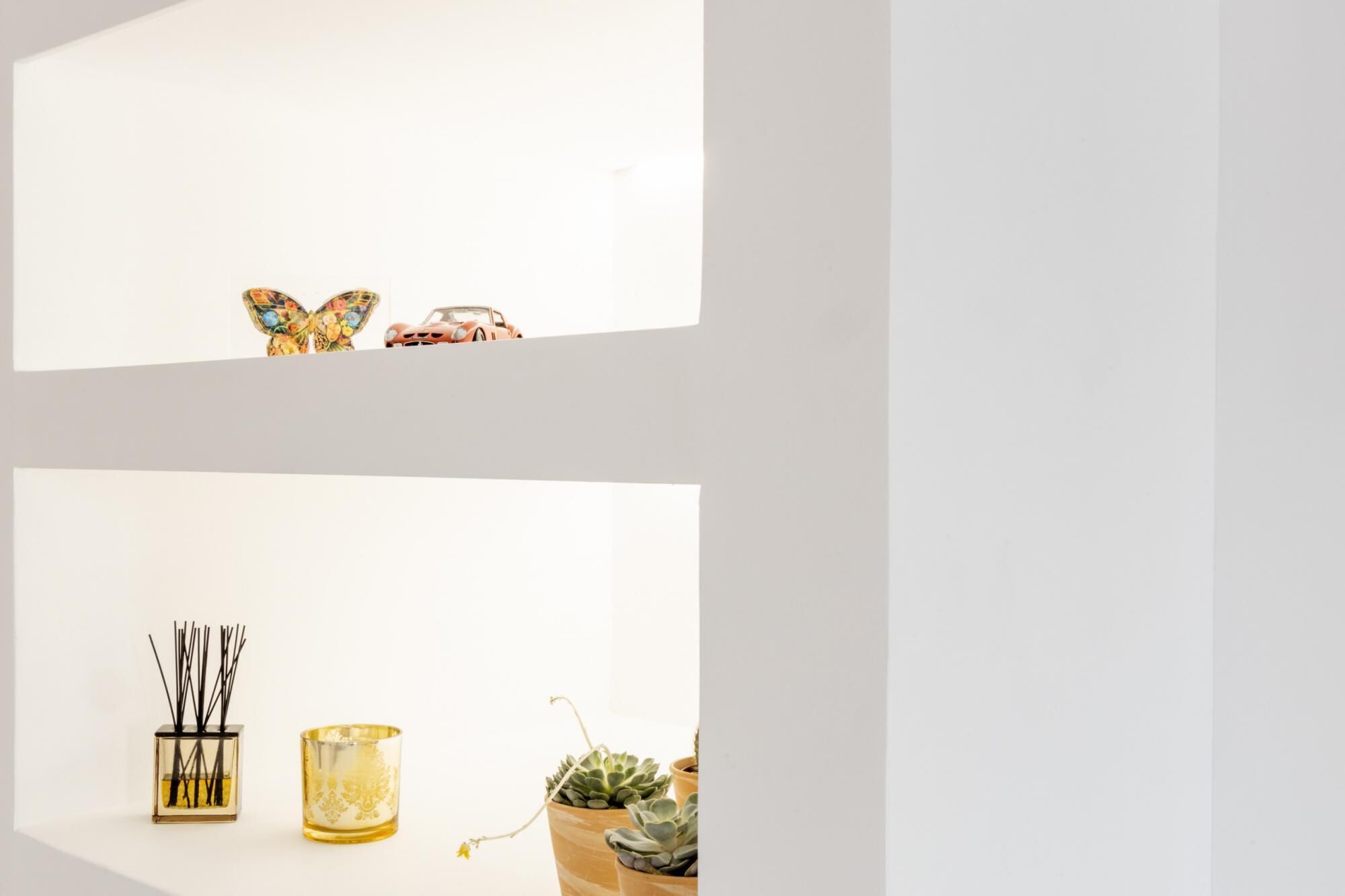 A minimalist decor with a butterfly and a car model on a shelf, a glass container with reeds, a candle, and a potted succulent.