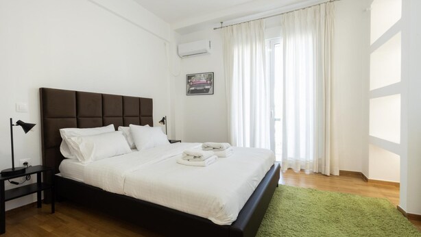 A neatly made bed with white linens and two towels, a bedside table with a lamp, and a green rug on a wooden floor. - Beautiful 2 Bdrm apt in Athens (Athens)