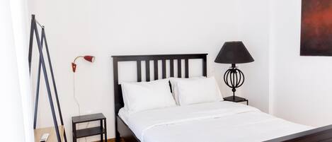 Una cama bien hecha con sábanas blancas, un cabecero de madera oscura, una mesita de noche con una lámpara y un pequeño estante con un dispositivo.