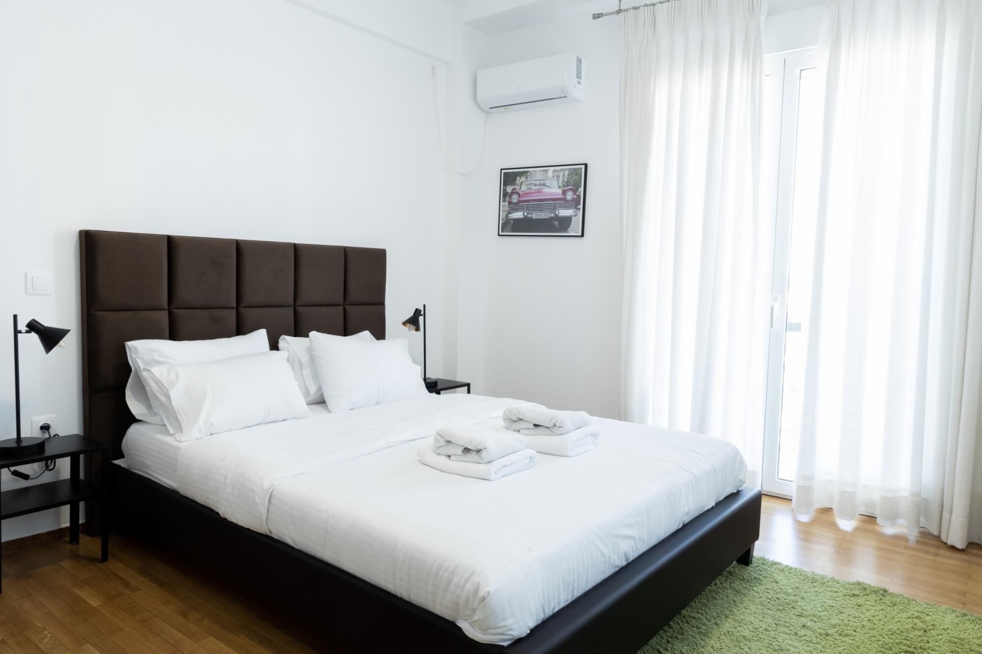 A neatly made bed with white linens and a dark headboard in a bright room with a green rug.