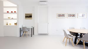 A modern, minimalist room with a white color scheme, featuring a dining area with a round table and chairs, a white radiator, and a wall-mounted air conditioner. - Beautiful 2 Bdrm apt in Athens (Athens)