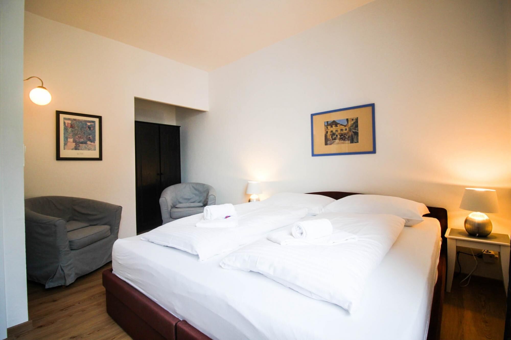 Een hotelkamer met een bed, een stoel, een bureau met een lamp, en twee ingelijste foto's aan de muur.