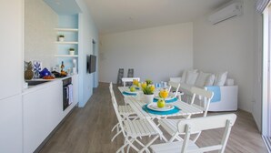 Una cocina moderna con armarios blancos, un comedor con sillas y una mesa dispuesta para cuatro, y un sofá blanco en la sala de estar.