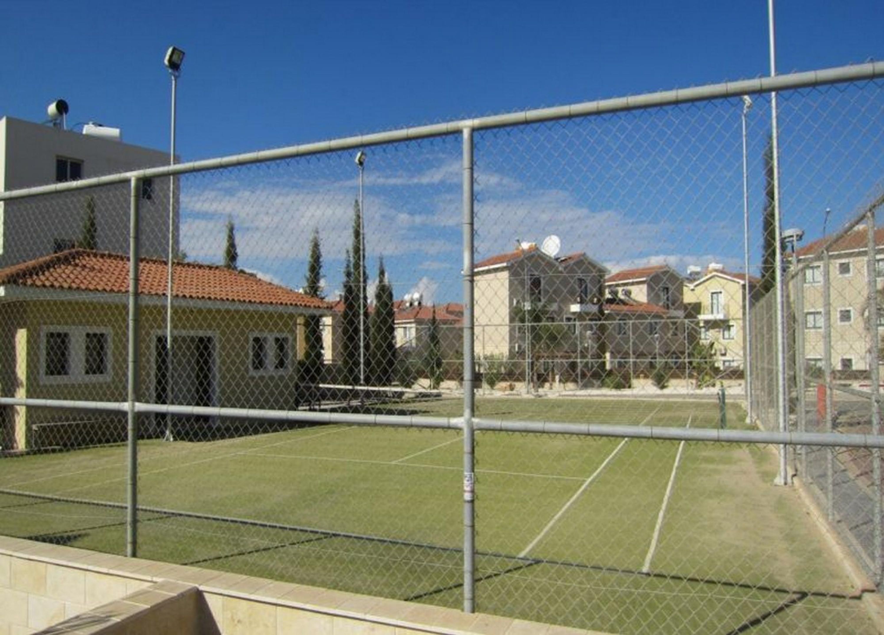 A fenced tennis court with a clear sky, surrounded by residential buildings.