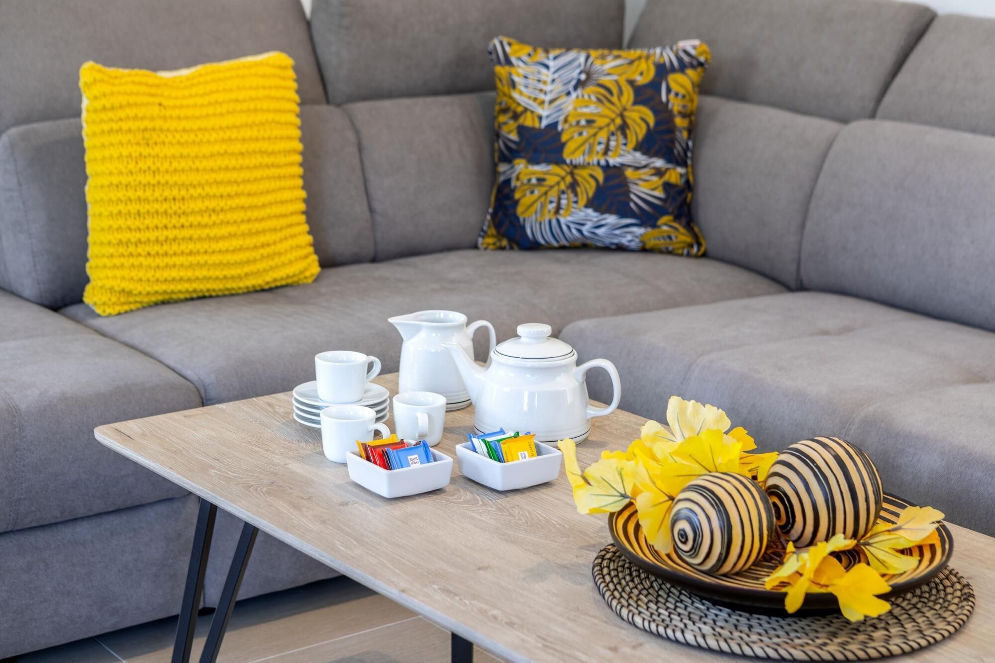 Una sala de estar con un sofá gris, una mesa de centro de madera y un cuenco decorativo con flores amarillas.