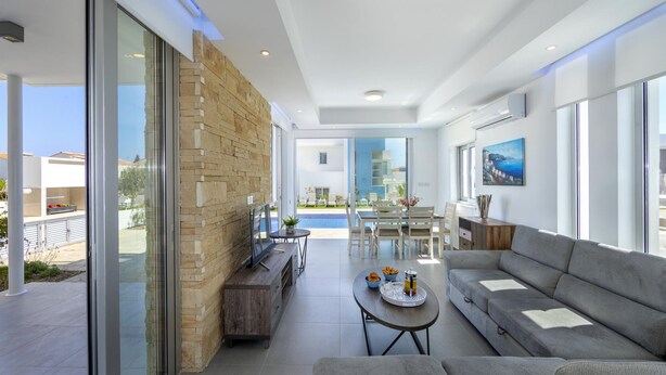 A modern living room with a stone wall, a sofa, a coffee table, and a dining area with a pool view. - Protaras Villa Mylos 9 (Paralimni)
