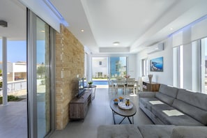 A modern living room with a stone wall, a sofa, a coffee table, and a dining area with a pool view. - Protaras Villa Mylos 9 (Paralimni)