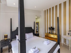 Uma cama com dossel com lençóis brancos e um edredom acolchoado, uma mesinha de cabeceira de madeira com abajur, um penteou dourado com espelho e uma cadeira branca com almofada.