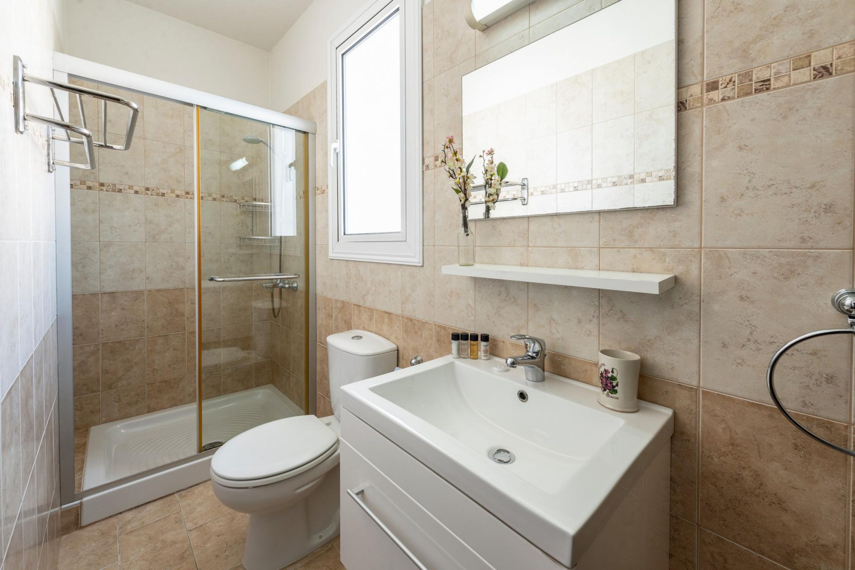 Een moderne badkamer met een glazen douchecabine, een wit toilet, een grote wastafel met een spiegel erboven, en een klein plankje met toiletartikelen.