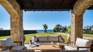A covered outdoor seating area with a view of the sea, stone pillars, and a wooden table with cushions and a book. - Palmrise Luxury Villas - Blue Villa (Kassandra)