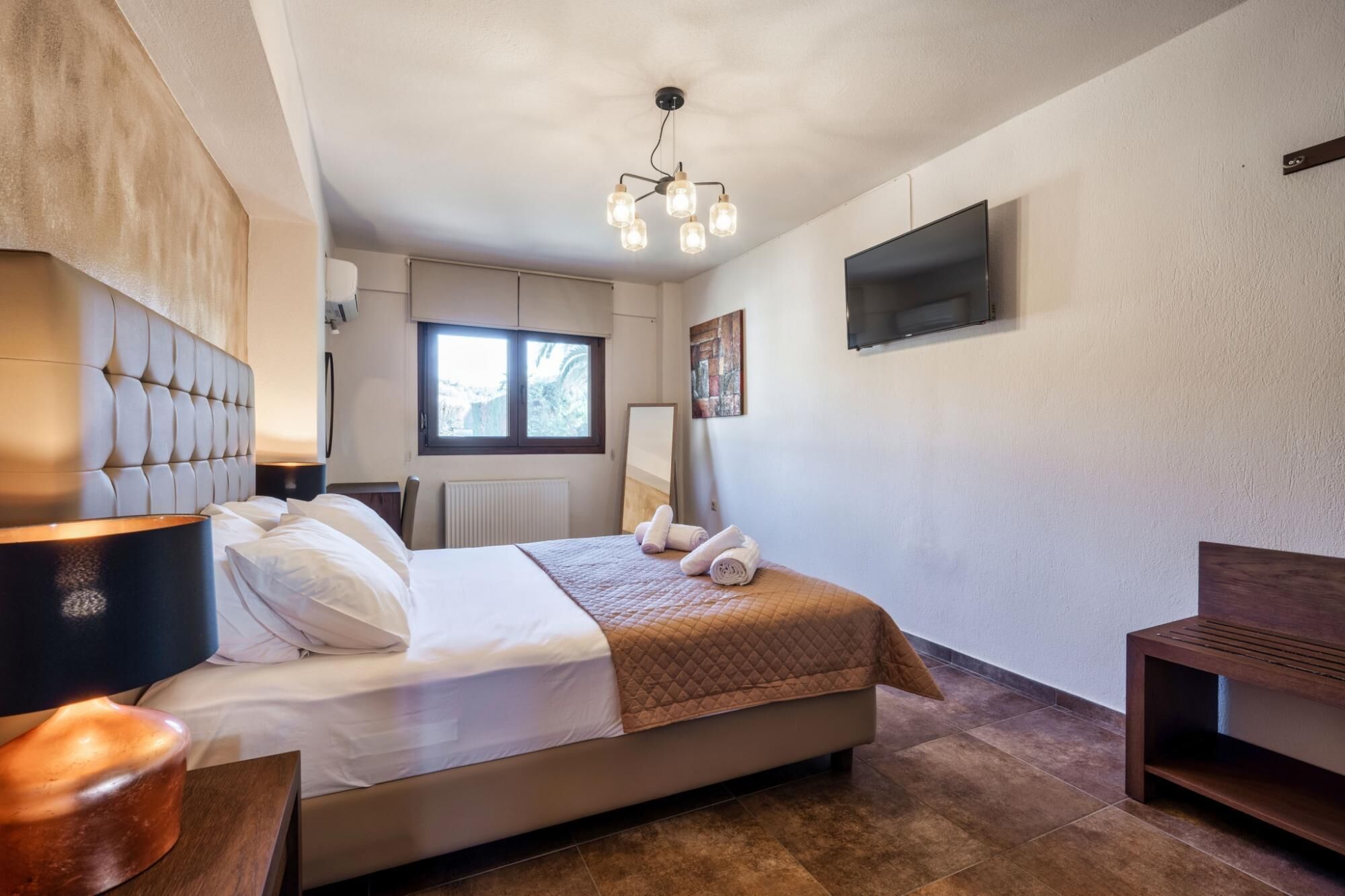 A hotel room with a bed, bedside lamp, artwork, a flat-screen TV, and a window.