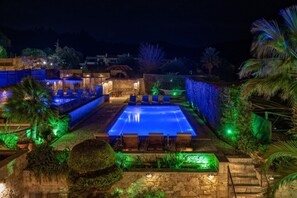 A nighttime swimming pool with lounge chairs and stone walls, surrounded by greenery and illuminated by blue and green lights.