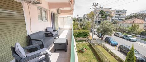 Un balcón con mobiliario de mimbre, una persiana cerrada y vista a una calle con coches aparcados y edificios.