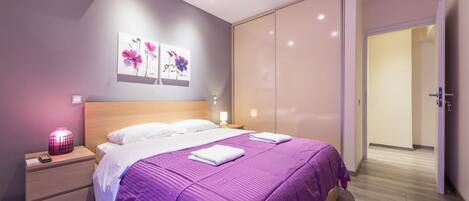 A modern bedroom with a bed, bedside tables, a nightstand, a wall-mounted air conditioner, and a door leading to another room.
