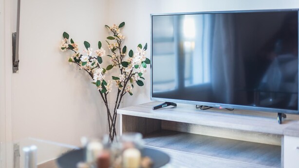 Ett modernt vardagsrum med en platt-TV på ett träställ, en vas med grenar och blommor och ett suddigt bord med diverse föremål.