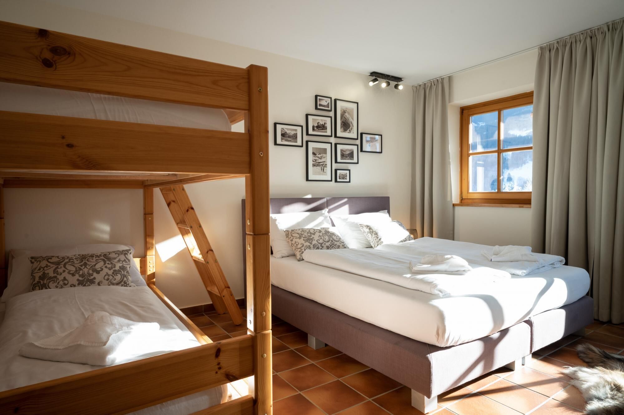 Ein Zimmer mit Etagenbett, einem Einzelbett und einem Fenster mit Vorhängen. An der Wand befinden sich gerahmte Bilder.