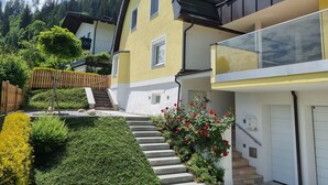 Una casa a due piani con un balcone, una scala che conduce all'ingresso e un giardino curato con siepi e fiori.