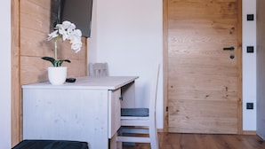 Une petite pièce moderne, dotée d’un bureau en bois, d’une chaise blanche, d’une orchidée en pot et d’un téléviseur à écran plat fixé au mur.