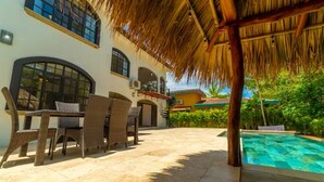 An outdoor patio with a table and chairs, a pool, and a building with large windows and a thatched roof. - Casa 48· Poolside Relaxing Retreat Near the Beach (Esterillos Este)