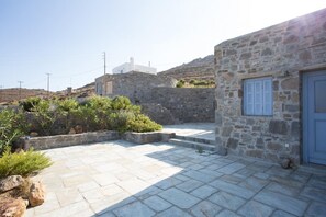 A stone courtyard with steps leading to a door, surrounded by greenery and a hillside in the background. - An Amazing Stone Villa-b in Serifos w Shared Pool (Serifos)