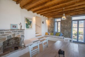 A room with a stone fireplace, wooden ceiling beams, a stone wall, a window with a view, and a seating area with cushions. - An Amazing Stone Villa-b in Serifos w Shared Pool (Serifos)