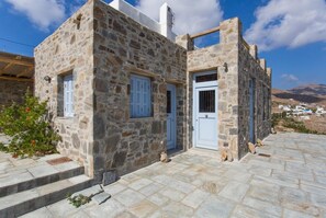 A stone building with a white door and shutters, a small porch, and a view of the landscape. - An Amazing Stone Villa-b in Serifos w Shared Pool (Serifos)