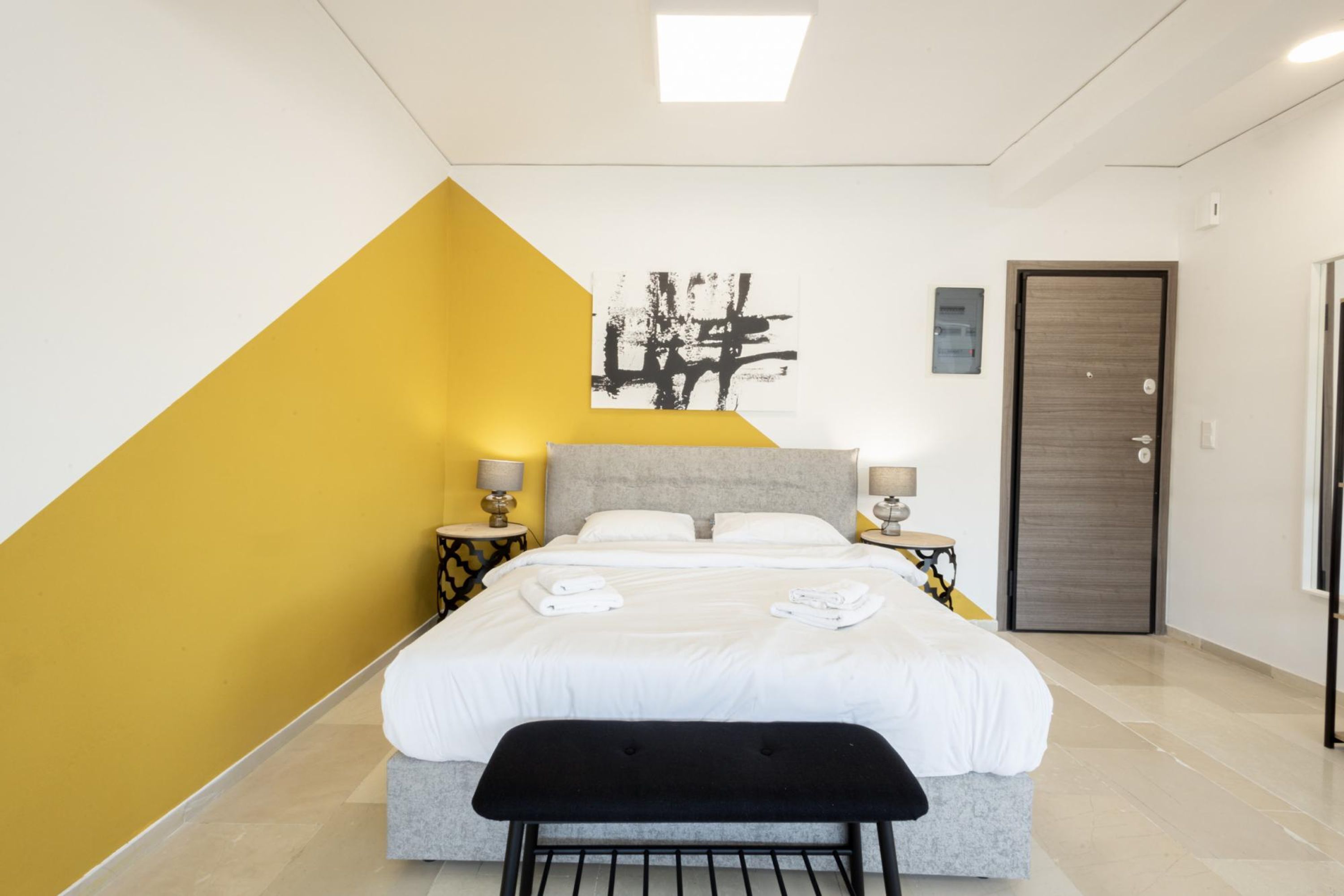 Een moderne hotelkamer met een bed, nachtkastjes, een bankje, een deur en een kunstwerk aan de muur.