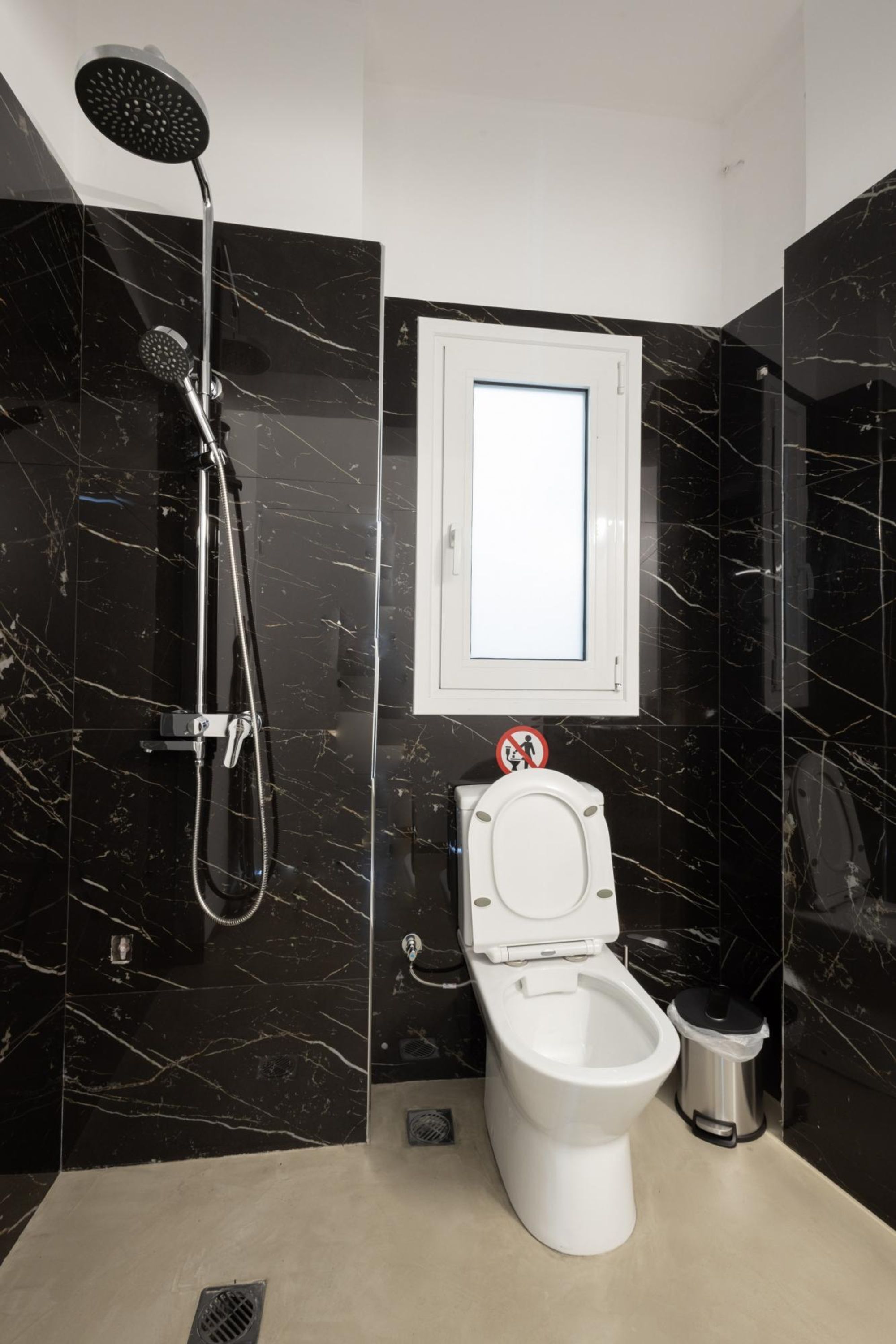 A modern bathroom with a black marble wall, a white toilet, a showerhead, and a trash bin.