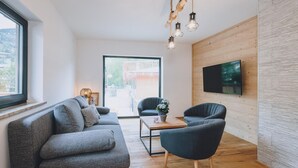 A modern living room with a grey sofa, a dark armchair, a wooden coffee table, and a TV mounted on a wooden wall. - Premium Apartments Areit - Balcony (Zell am See)