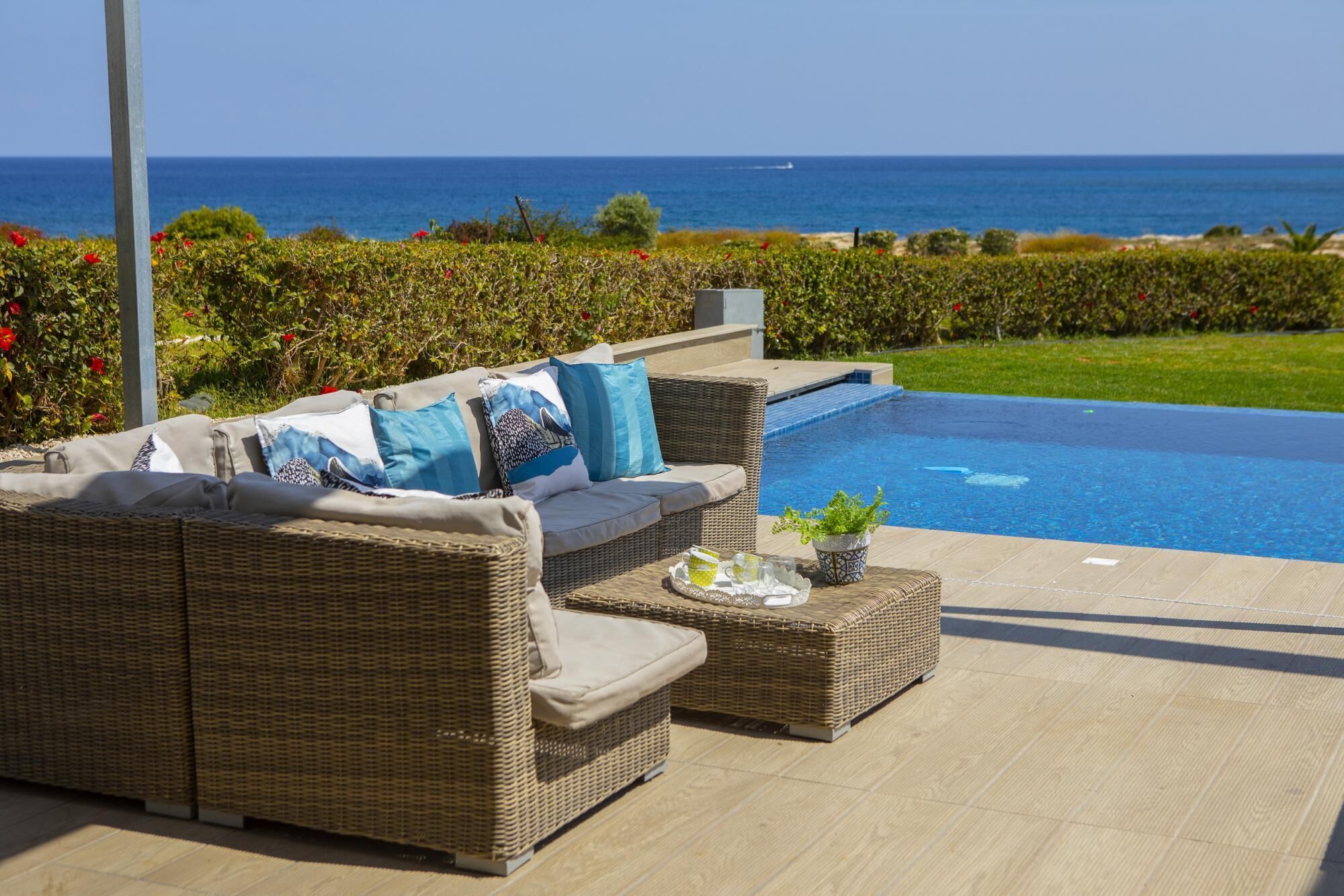 A wicker sofa with cushions and a tray of drinks by a pool with a view of the ocean.