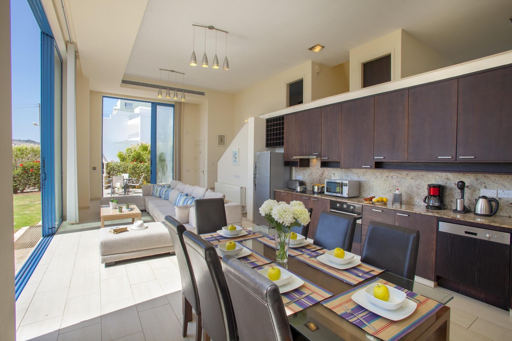 A modern kitchen with dark wooden cabinets, a built-in microwave, and a coffee maker. There is a dining area with a glass table and chairs, and a view of a garden through large windows.