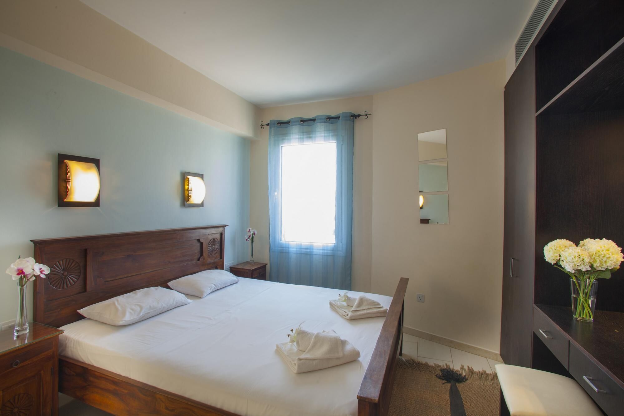 A hotel room with a large bed, two wall-mounted lights, a window with curtains, a mirror, and a dresser with a vase of flowers.