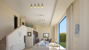 A spacious living area with a large sofa, a coffee table, and a dining table. There is a staircase leading to an upper floor, and a kitchen area is visible in the background. - Villa Infinite Aretousa (Paralimni)