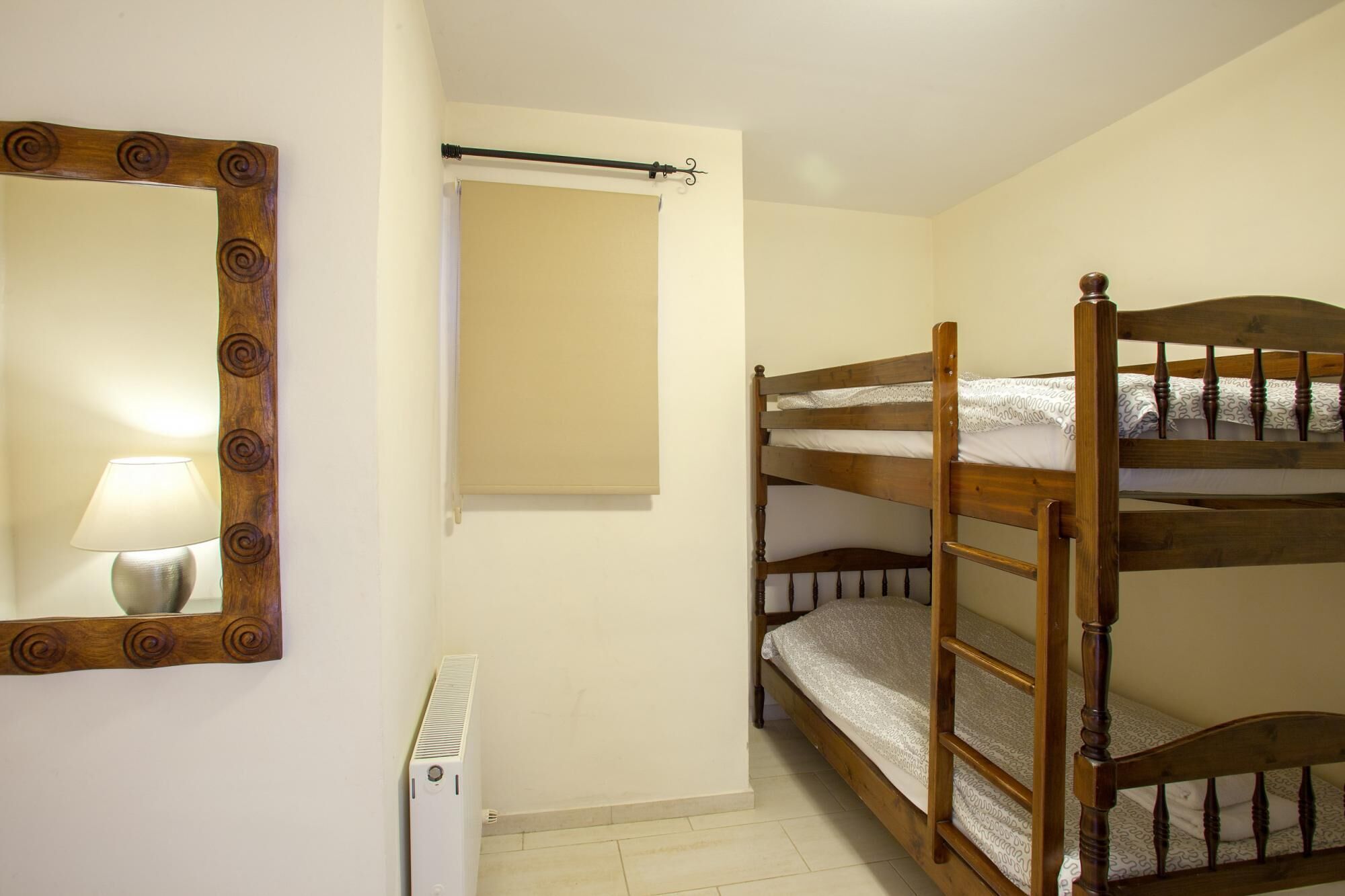 A small, single-bedded room with a wooden bunk bed, a bedside table with a lamp, a radiator, and a mirror with a decorative frame.