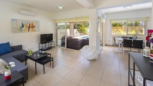 A spacious living area with a sofa, coffee table, and TV. There is a dining area with chairs and a table. The room has large windows and a view of the outdoors.