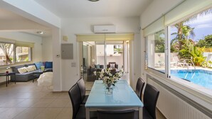 A dining area with a glass table and chairs, a view of a swimming pool, and a sofa in the background.