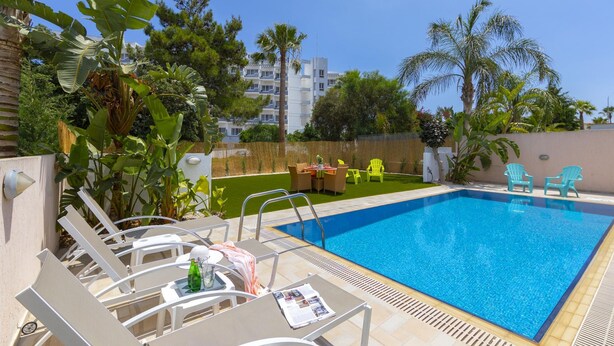 A swimming pool with lounge chairs and a table, surrounded by tropical plants and a clear sky.