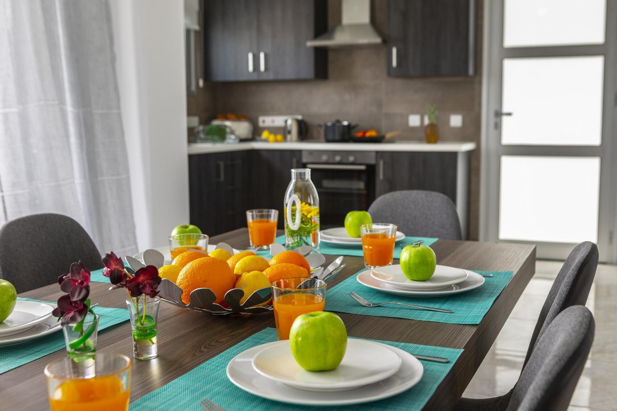 A dining table set with plates, cutlery, and fruits, with a kitchen in the background.