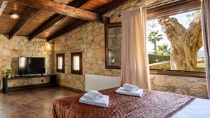 A bedroom with stone walls, a wooden ceiling, a large window with a view of palm trees, a bed with towels, and a TV on a console table. - Palmrise Luxury Villas - Thea Villa (Kassandra)