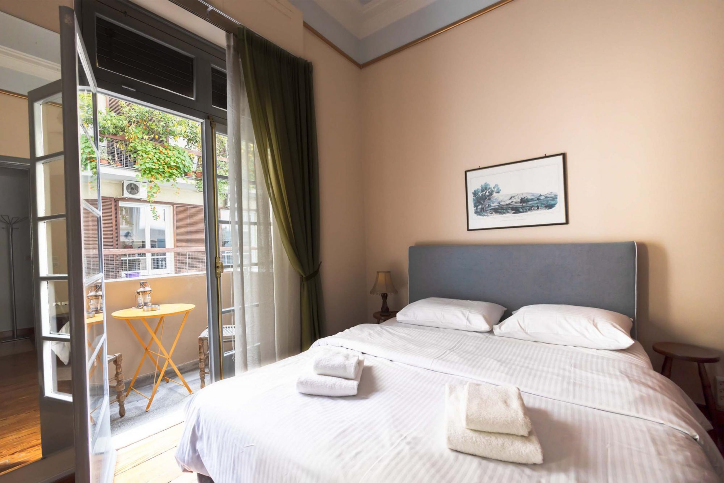 Hotelkamer met een bed, een kleintje tafeltje, een stoel, een raam met uitzicht op een gebouw en een foto aan de muur.