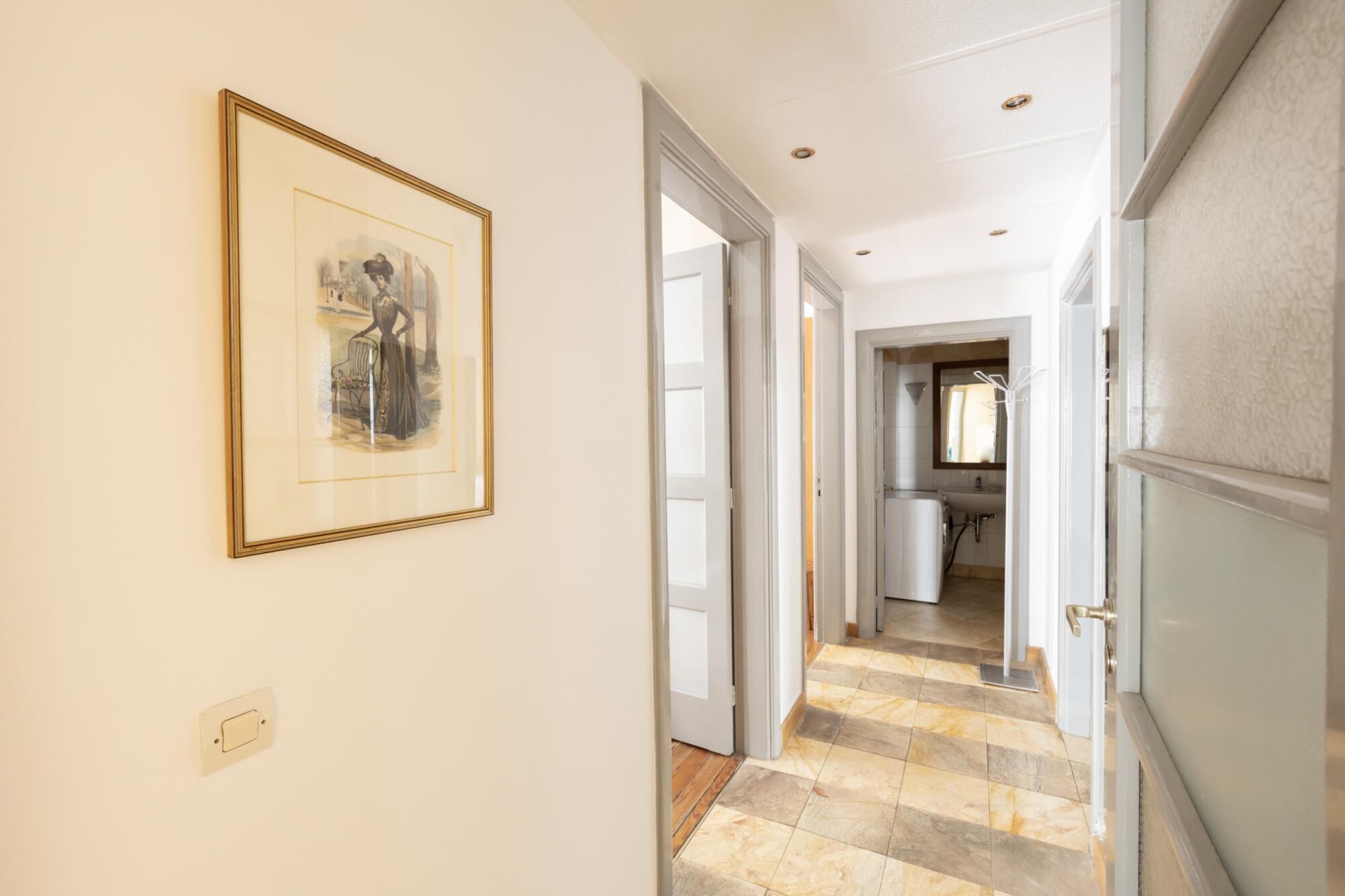 Un couloir orné d’un tableau encadré au mur, qui mène à une salle de bain équipée d’un lavabo et d’un miroir.