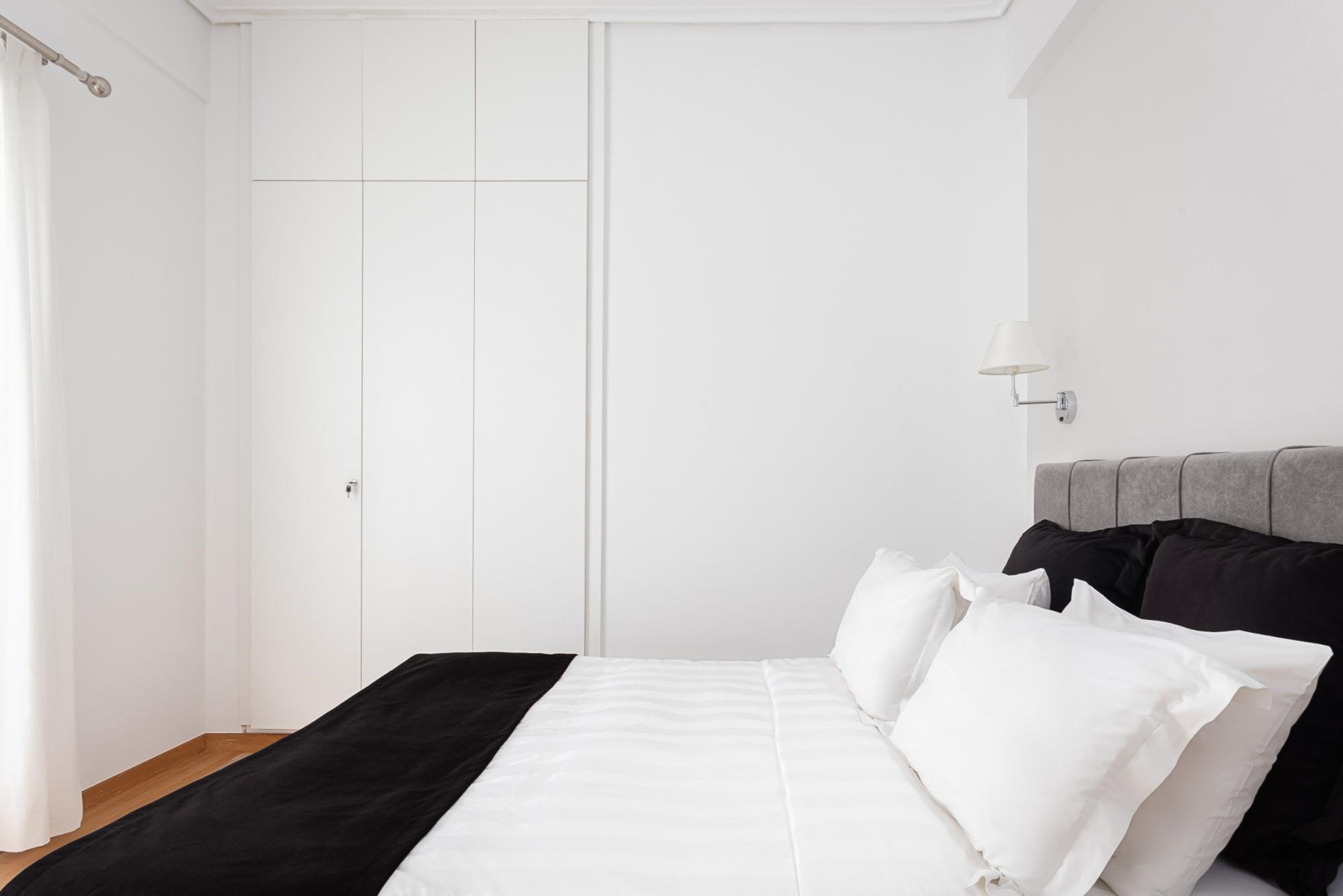 A bedroom with a bed, white curtains, a wardrobe, and a wall-mounted lamp.