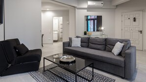 A modern living room with a grey sofa, black armchair, and a glass coffee table. There is a patterned rug on the floor, and a door with a sign is visible in the background. - 85 sqm in the Heart of Athens w Rooftop Deck (Athens)