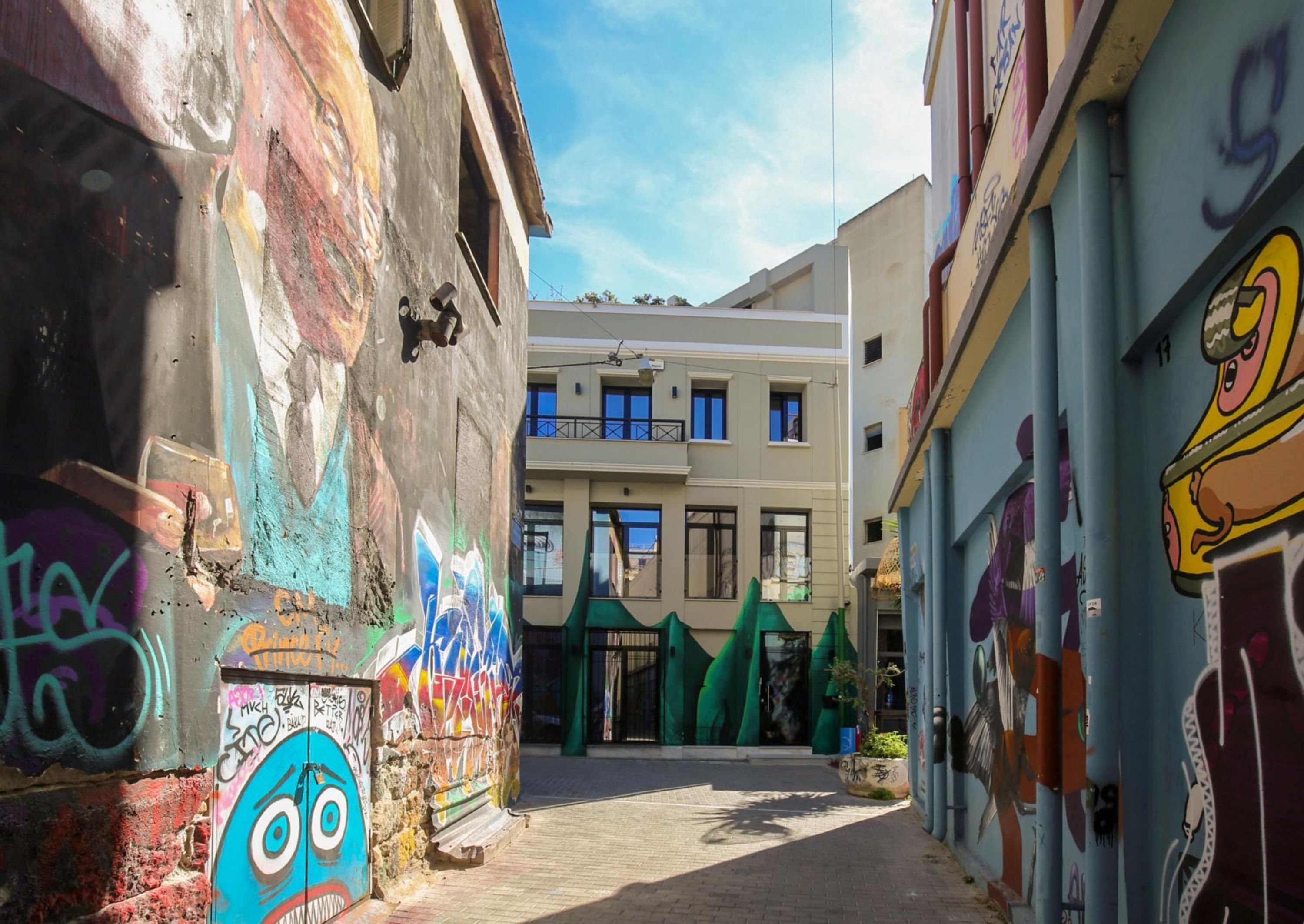 A narrow alley with colorful graffiti on the walls, leading to a building with a modern facade and large windows.